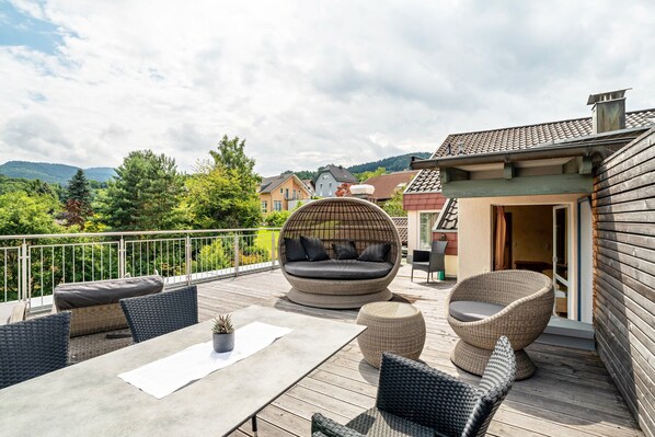 Une terrasse en bois, aménagée avec du mobilier d’extérieur, notamment un canapé et des fauteuils, qui offre une vue panoramique sur un paysage pittoresque, avec des maisons et des arbres.