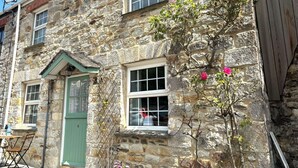 Un bâtiment en pierre avec une porte et des fenêtres vertes, un petit espace extérieur aménagé avec des sièges et une plante grimpante aux fleurs roses.