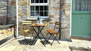 Un bâtiment en pierre avec une fenêtre et une porte verte. On y voit une table avec des chaises et une bouteille posée dessus.