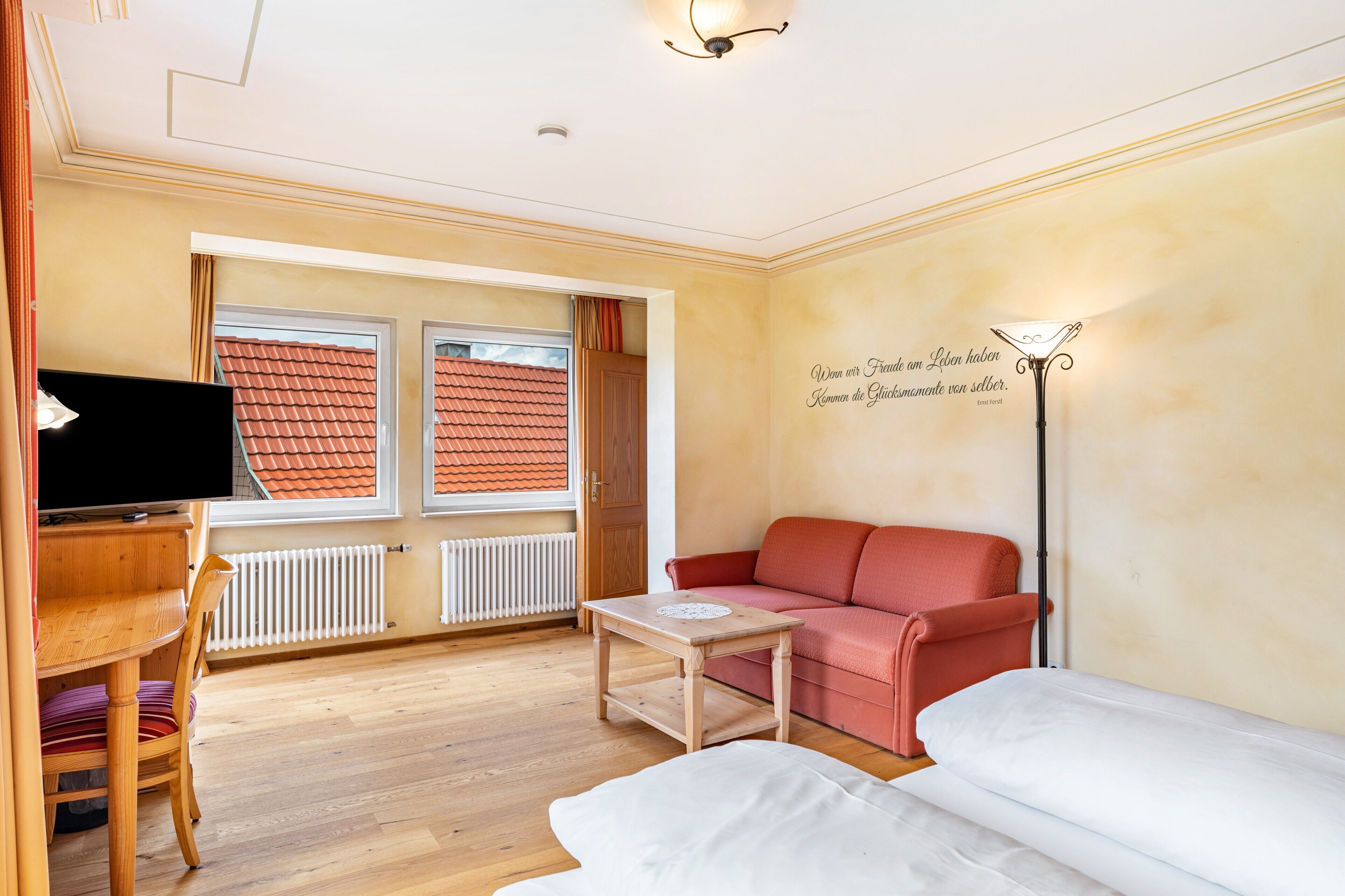 A living room with a sofa, a table, a TV, and a window with a view of a red-roofed building.