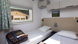 Una habitación pequeña con cama individual, ventana, ropa de cama blanca, manta colorida, cabecera y estante fijado a la pared.