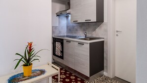 Eine kompakte Küche mit grauen Schränken, einer Edelstahl-Spüle und einem kleinen Backofen. Auf einem Tisch steht ein Topf mit Pflanze und an der Wand hängt eine Uhr.