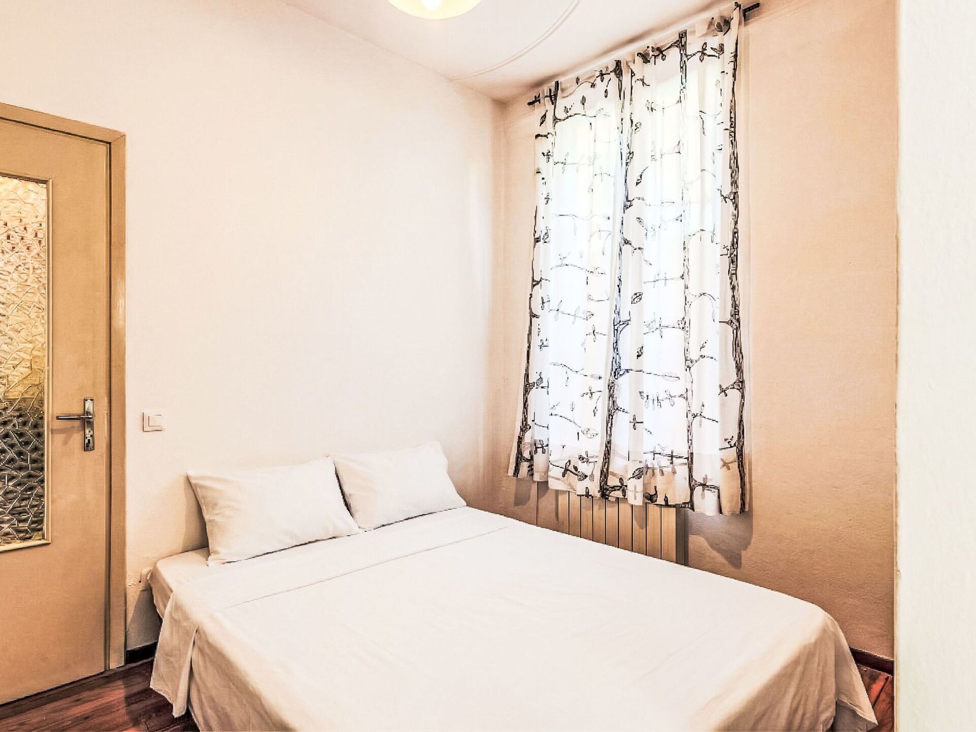 Un dormitorio con una cama, una puerta con celosía metálica y una ventana con cortinas estampadas.