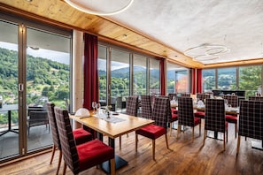 Restaurante com uma grande mesa e cadeiras, amplas janelas com vista para uma área arborizada e teto de madeira.