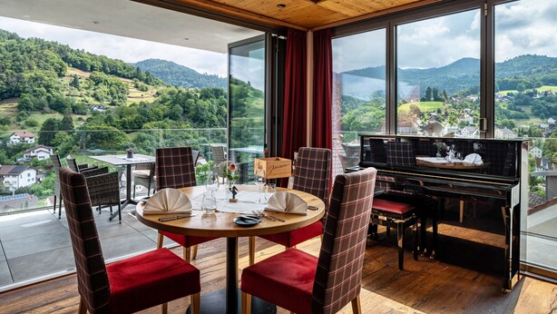 A dining area with a piano, red upholstered chairs, a round table, and a scenic mountain view through large windows.