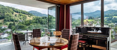 A dining area with a piano, red upholstered chairs, a round table, and a scenic mountain view through large windows.