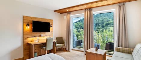 Une chambre d’hôtel comprenant un lit, un bureau avec une chaise, un téléviseur fixé à un mur en bois et un balcon donnant sur un paysage verdoyant.
