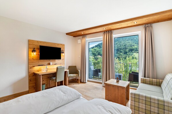 Habitación de hotel con cama, escritorio con silla, televisor montado en una pared de madera y balcón con vista a zonas verdes.