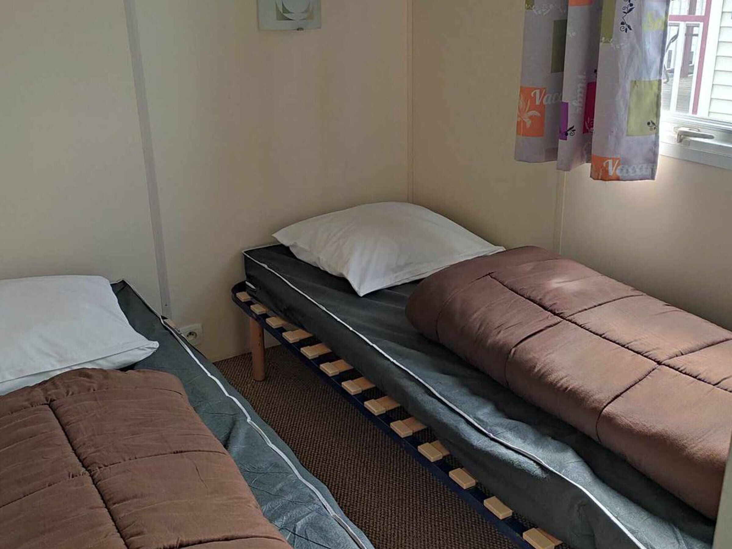 A dormitory room with two single beds, a window with curtains, and a small wall-mounted light fixture.