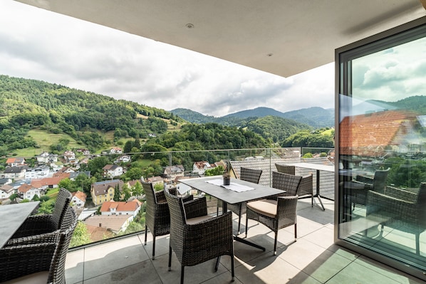 Un balcon doté de meubles en osier, d’une table et d’une porte vitrée donnant sur une ville et des montagnes.