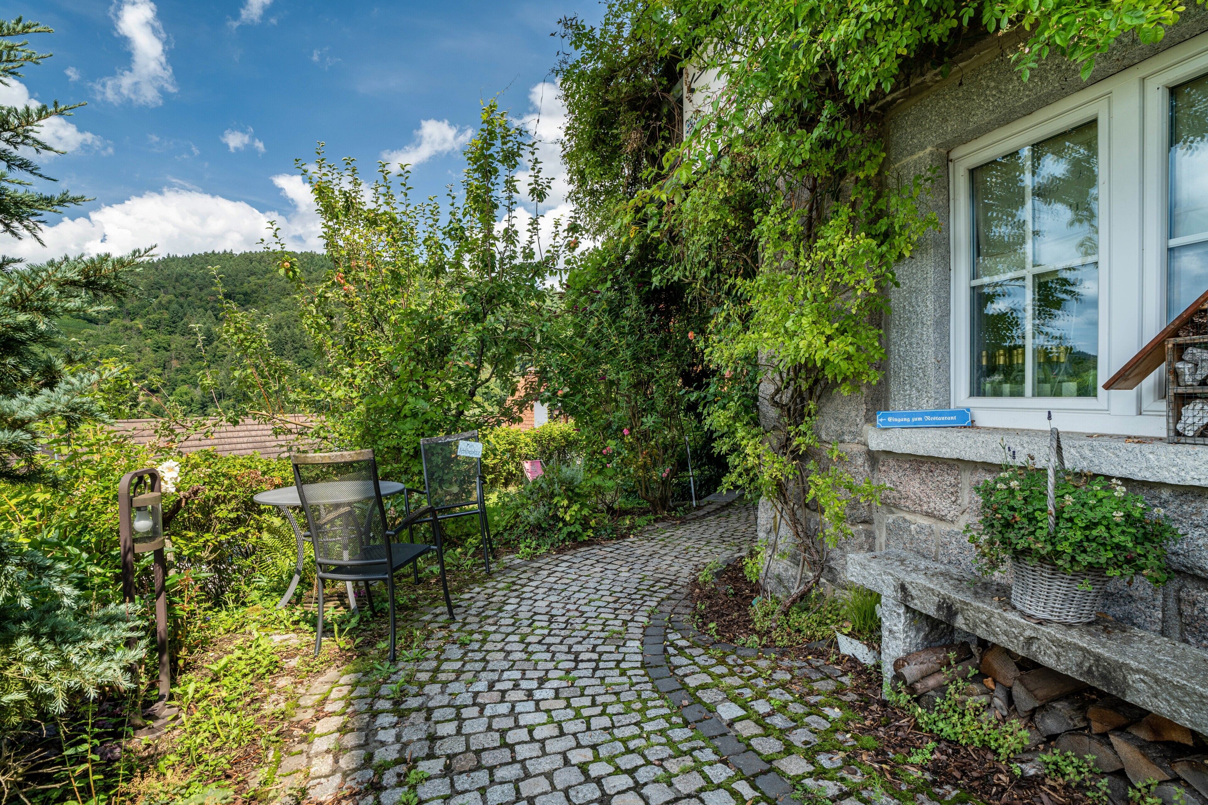 Une terrasse en pierre avec une table et des chaises, entourée de verdure et d’un mur de pierre.