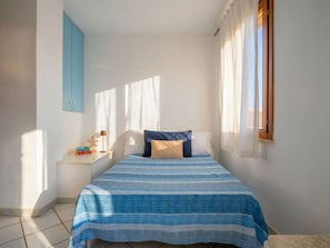 A small, bright bedroom with a single bed, a nightstand, a window with curtains, and a blue wall.
