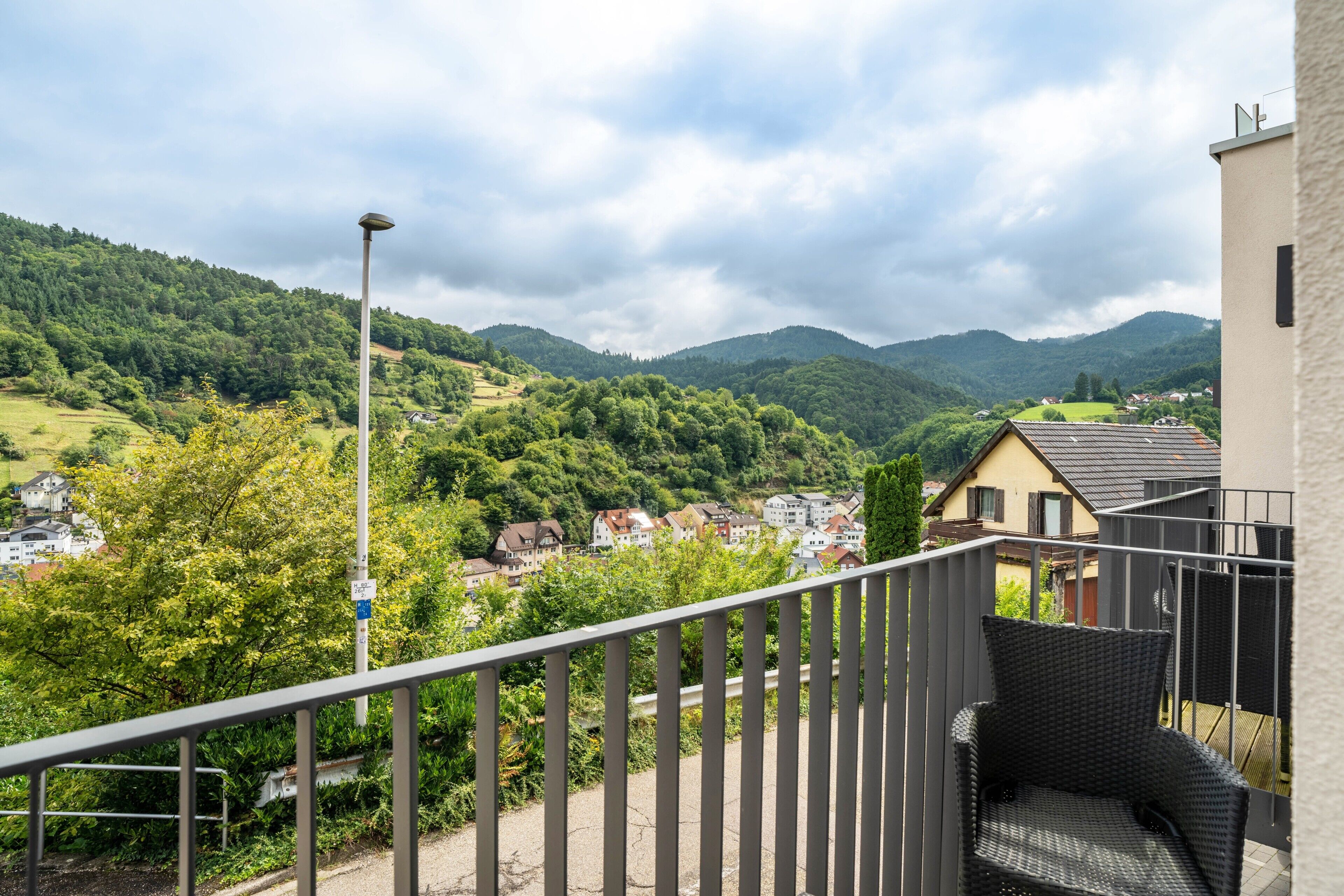 A balcony with a wicker chair, a street lamp, and a view of a town with houses and green hills in the background.