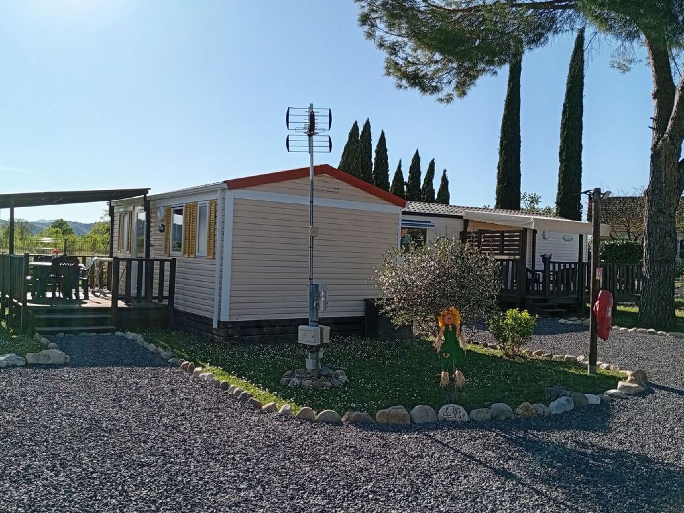 Una casa móvil con techo rojo, un patio con mesa y sillas, y un pequeño jardín con un árbol y arbustos.