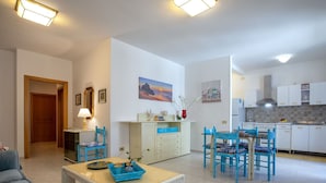 A living room with a sofa, a coffee table, and a dining area with chairs and a table. There is a kitchen with cabinets and a stove. - Nice apartment for 5 guests with A/C, WIFI, TV and terrace (Alghero)
