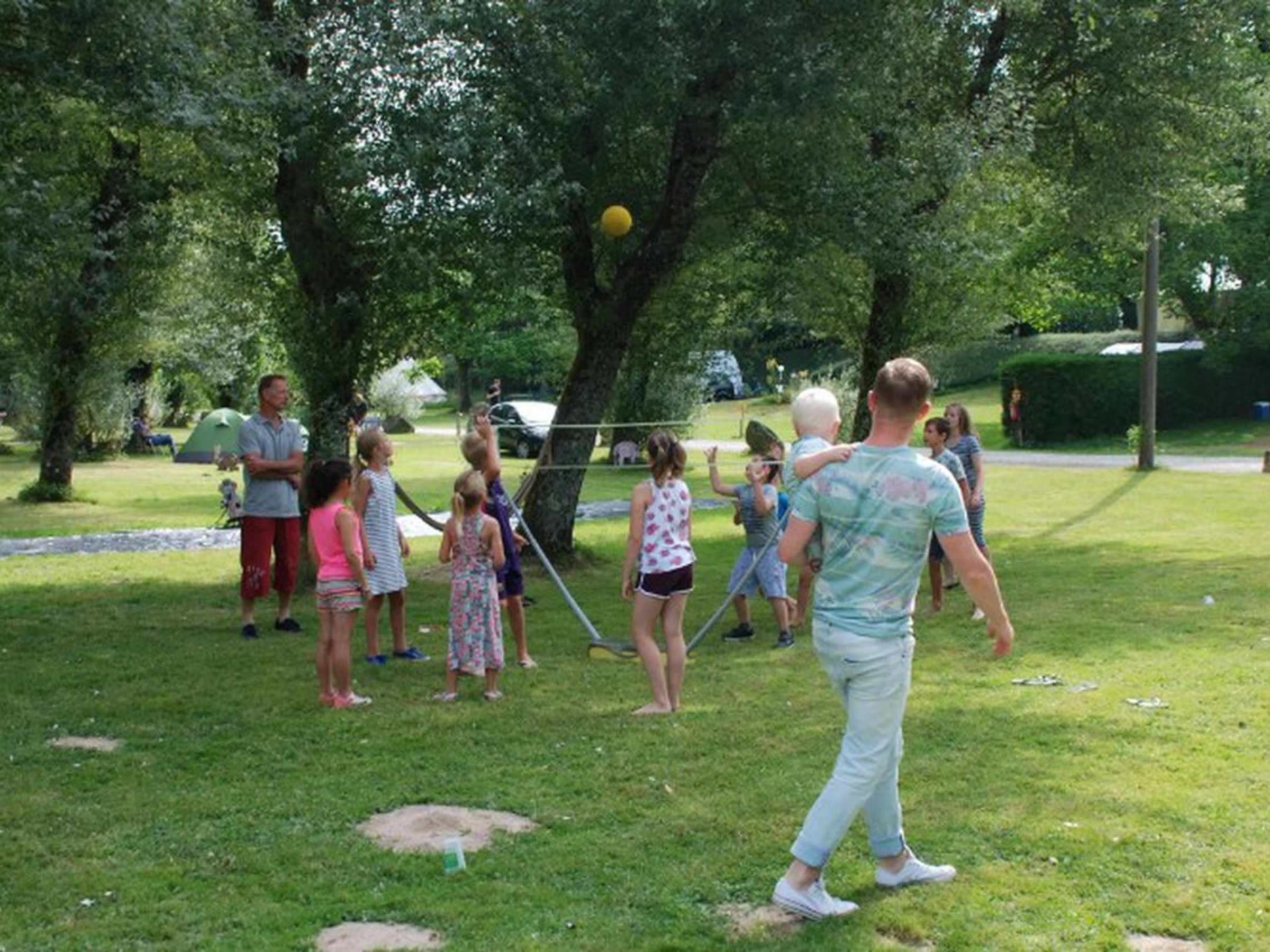 Un groupe de personnes joue avec un ballon jaune dans un parc verdoyant, avec des arbres et une tente en arrière-plan.
