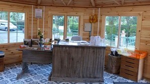 A wooden cabin interior with a central wooden desk, a trash can, and a window with a view of a car and greenery outside.