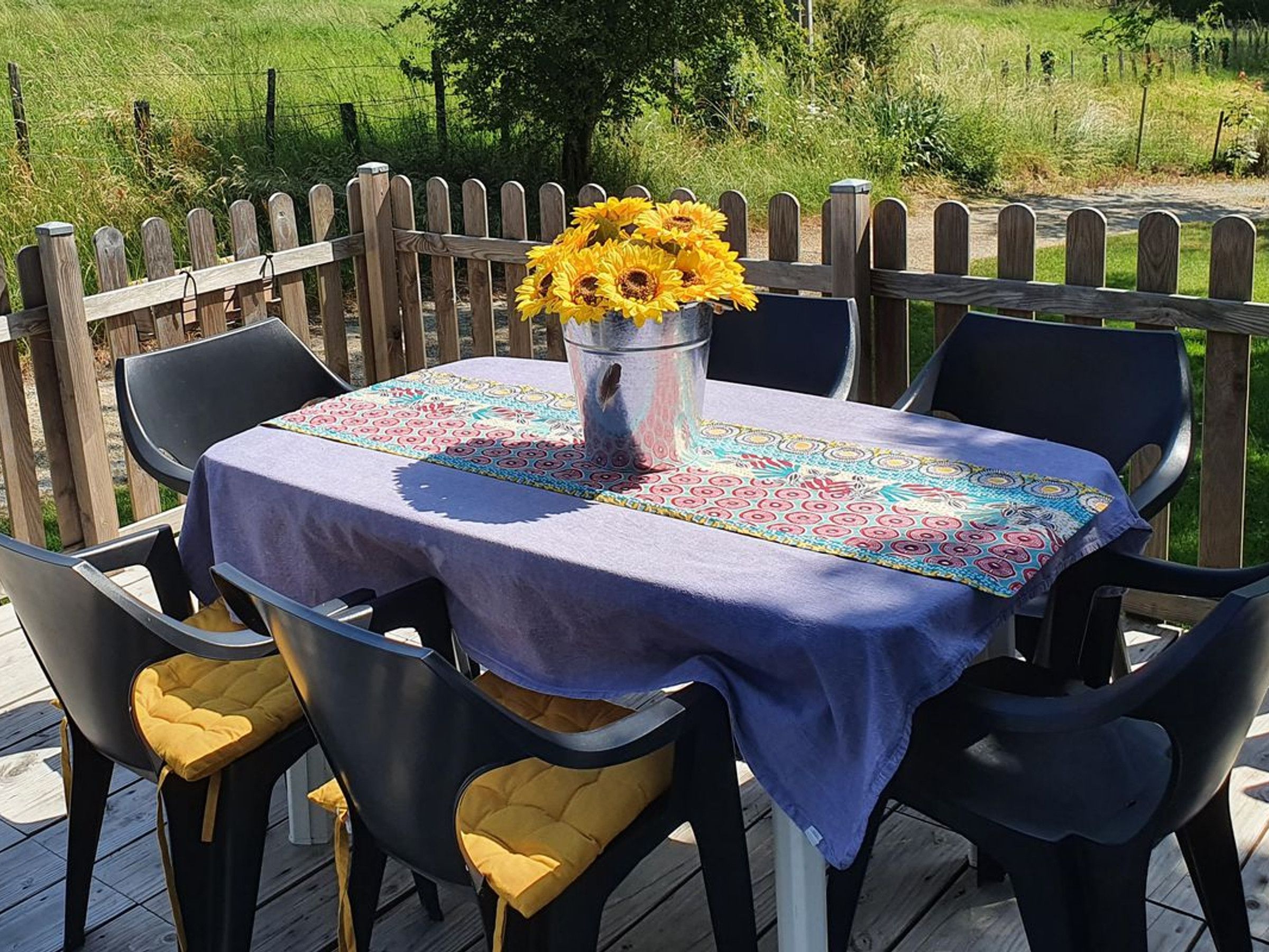 Un espace de restauration en plein air avec une table dressée pour deux personnes, entourée de chaises dotées de coussins jaunes, d’une nappe décorative et d’un vase rempli de fleurs jaunes.