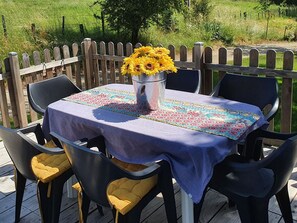 Un área de comedor al aire libre con una mesa dispuesta para dos, rodeada de sillas con cojines amarillos, un mantel decorativo y un jarrón con flores amarillas.