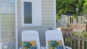 Deux chaises longues en plastique blanc, ornées de coussins aux motifs tropicaux, sont posées sur une terrasse en bois.