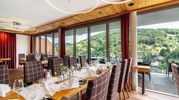 A dining area with a long table set for a meal, large windows offering a view of a forested landscape, and a wooden ceiling.