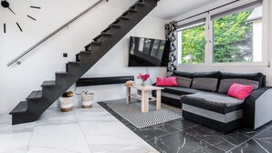 Ein modernes Wohnzimmer mit einer schwarzen Wendeltreppe, einem grauen Sofa mit rosafarbenen Kissen, einem Flachbildfernseher und einem großen Fenster mit Blick auf Grünflächen.