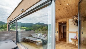 Een modern terras buiten met glazen wanden, houten vloer en uitzicht op een bergachtig landschap.