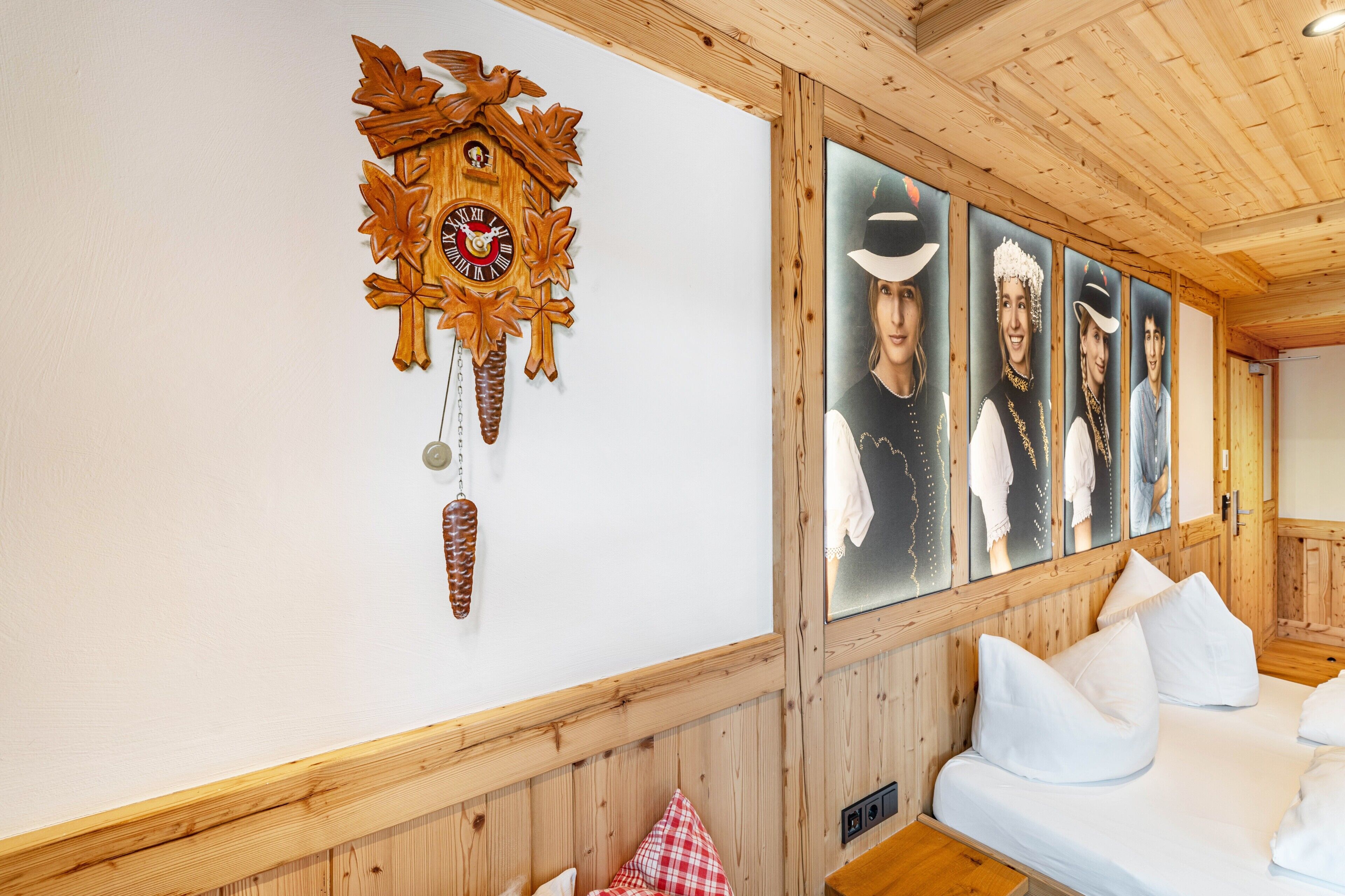 A wooden interior with a cuckoo clock, framed portraits, and a bed with white bedding.