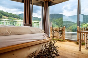 A bedroom with a large bed, wooden floor, and a view of green hills through large windows.