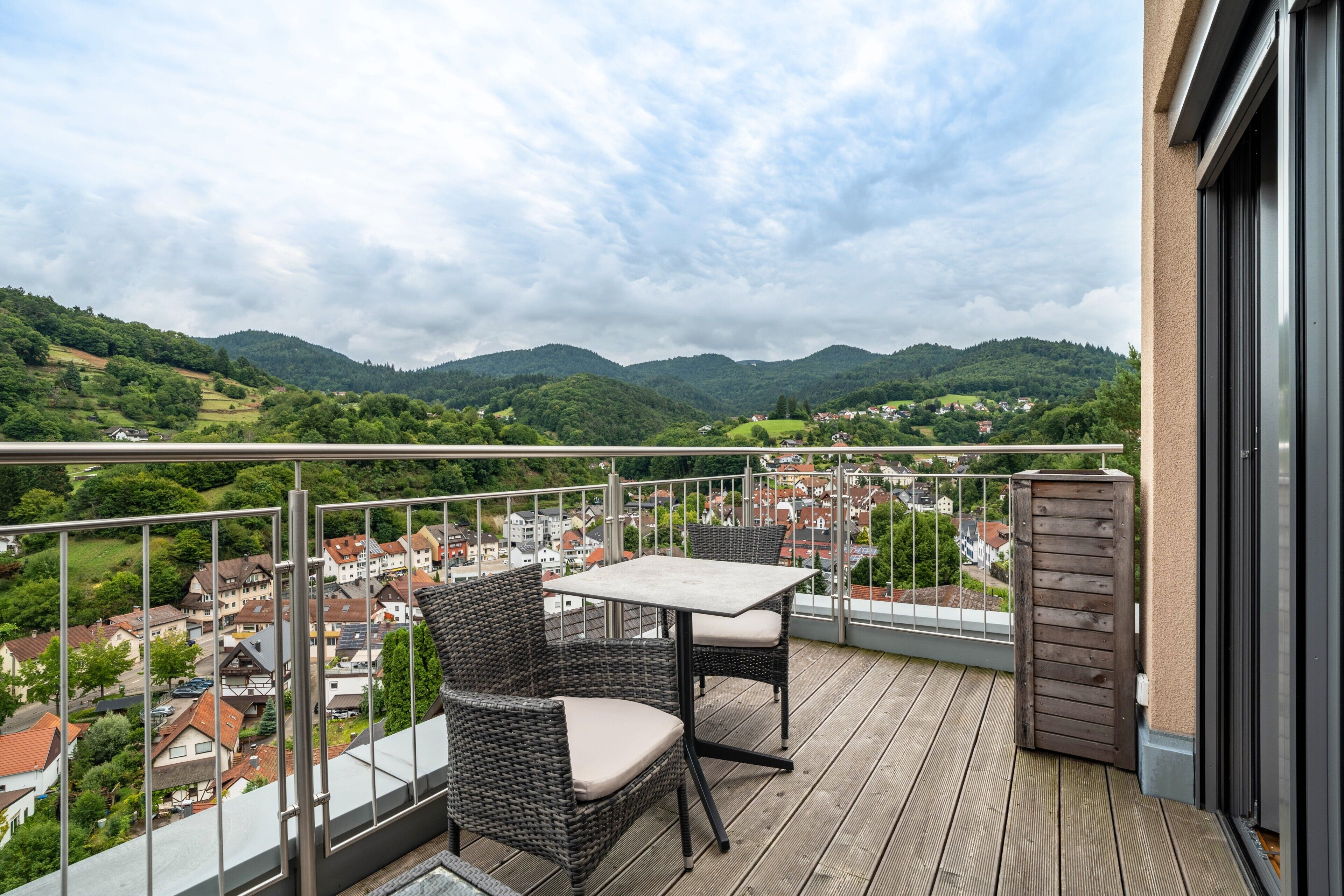 A balcony with wicker furniture, a table, and a view of a town and hills.