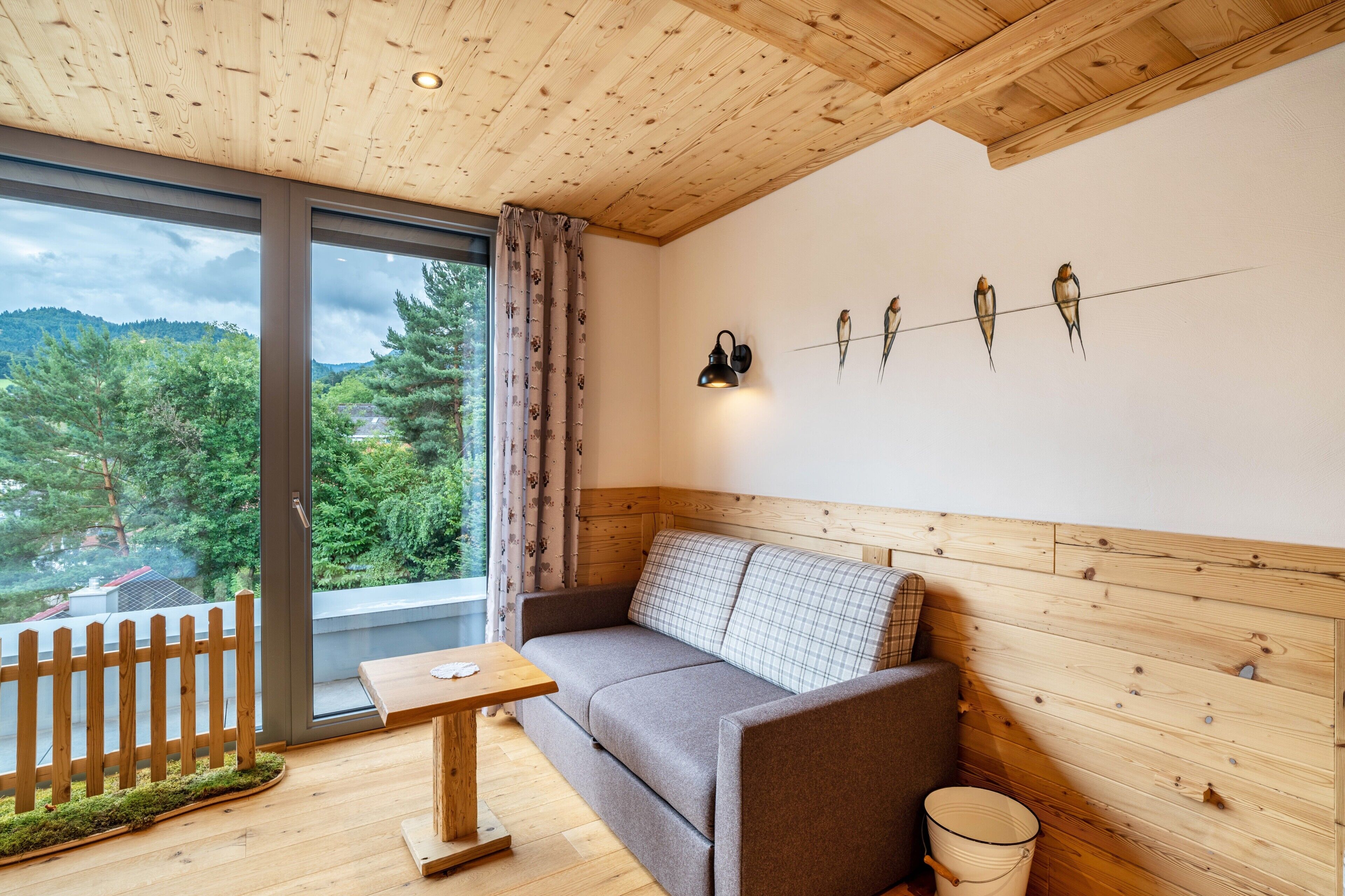 Una habitación con techo de madera, un sofá, una mesa, una lámpara fijada a la pared y una ventana con vistas a árboles y una casa.