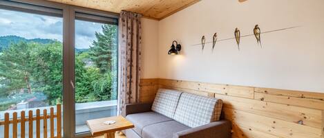 Una habitación con techo de madera, un sofá, una mesa, una lámpara fijada a la pared y una ventana con vistas a árboles y una casa.