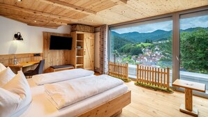 Ein Zimmer mit Holzboden, ausgestattet mit einem Bett, einem Schreibtisch, einem Fernseher und einem großen Fenster mit Blick auf eine Stadt und Berge.