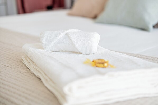 A neatly folded white towel with a yellow object on it, placed on a bed with a light-colored blanket and a beige pillow in the background.