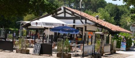 Café al aire libre con mesas y sillas bajo un toldo blanco, rodeado de follaje.