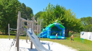 A playground with a slide and climbing structures, surrounded by trees and a blue inflatable play area.