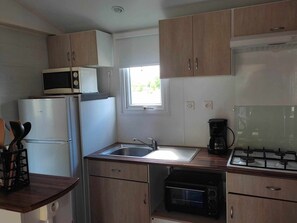 A compact kitchen with wooden cabinets, a microwave, a refrigerator, a sink, a stove, and a window providing natural light. - 32m² Mobile Home with Terrace for 4 People in Anduze (Anduze)