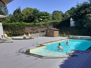 A swimming pool area with a wooden deck, a small wooden building, and lush greenery in the background.