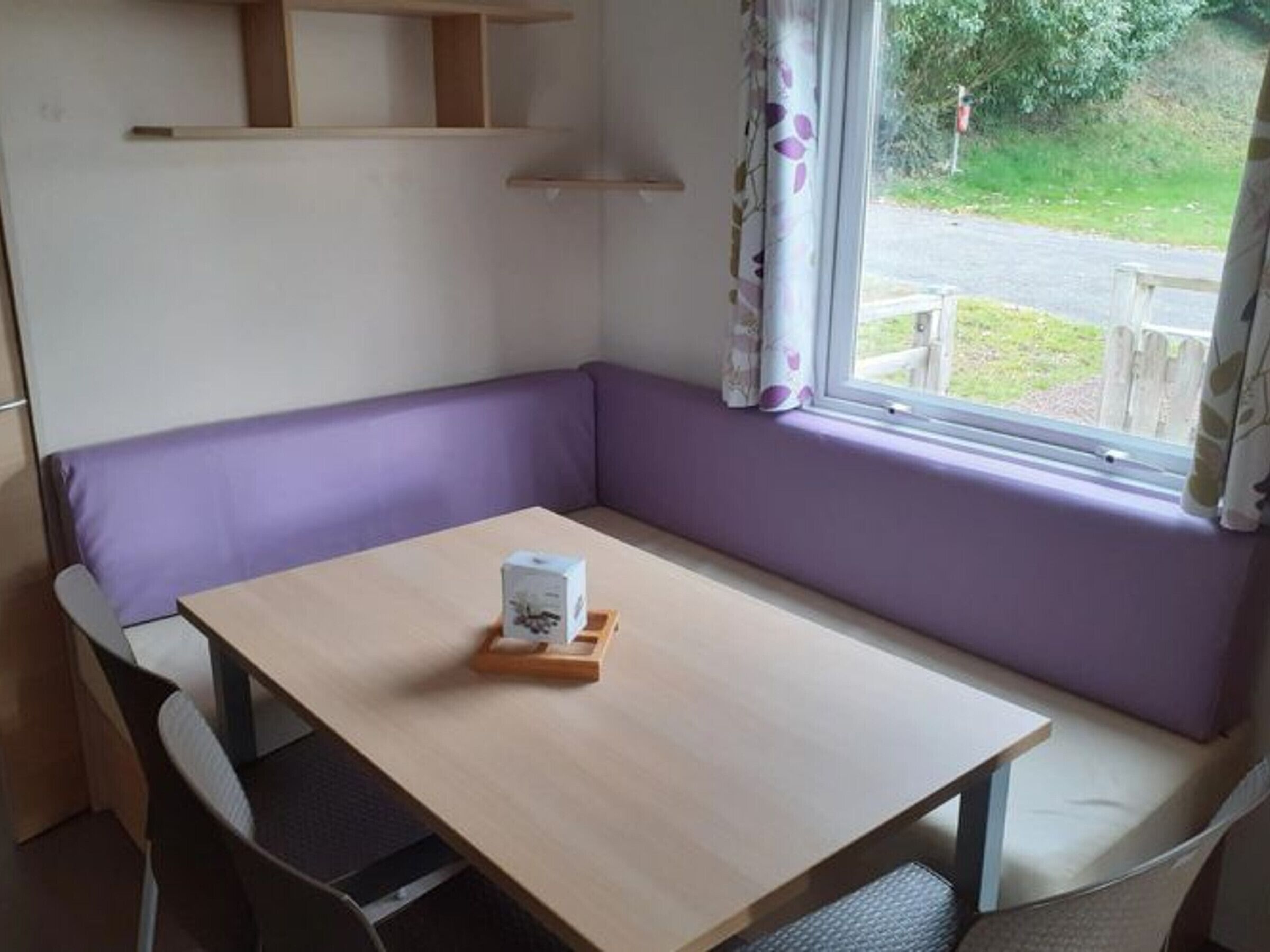 Habitación de esquina con mesa de madera, banca acolchada color púrpura y ventana con cortinas.