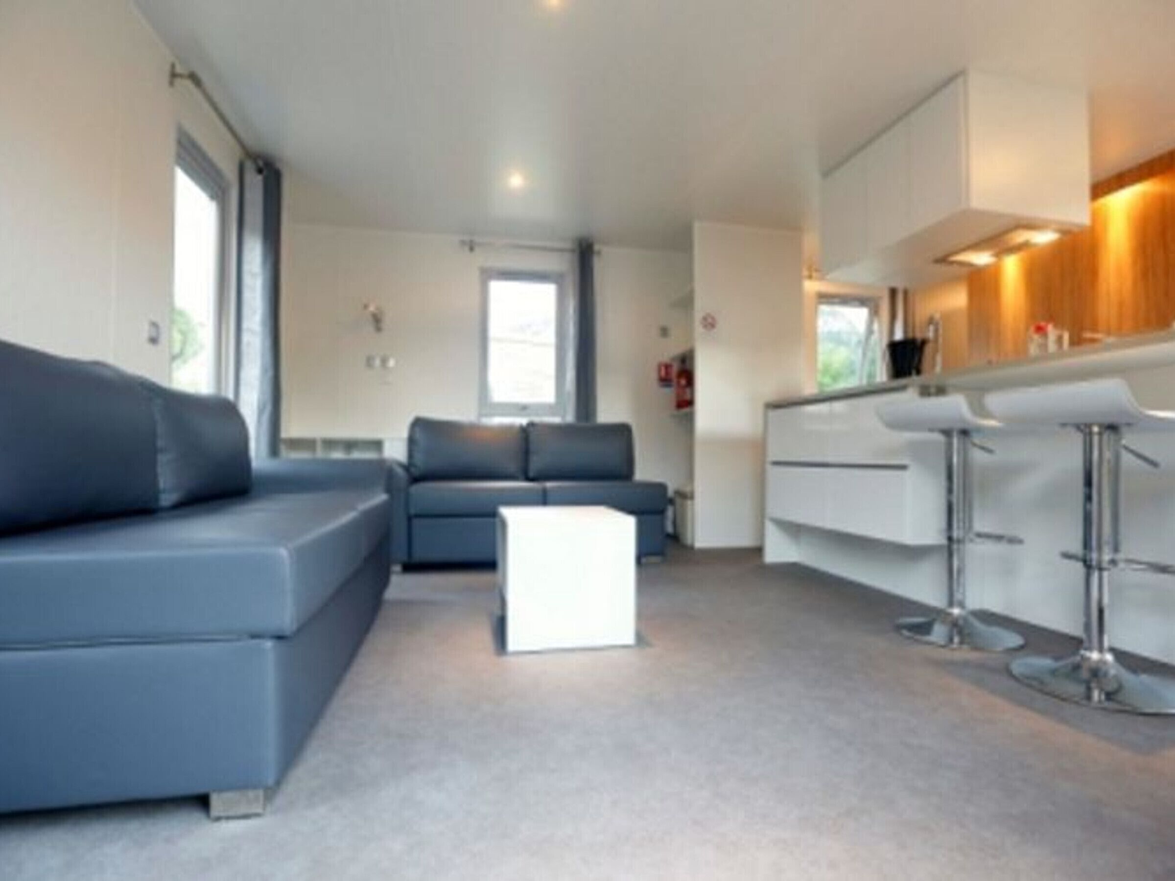 A modern living room with a grey sofa, a small white ottoman, and a kitchen area with white cabinets and bar stools.