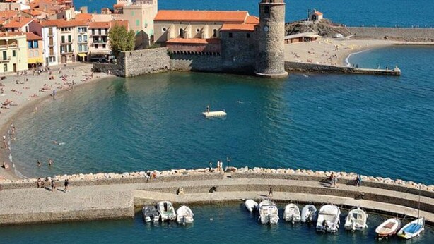 Uma cidade costeira com uma torre histórica, uma praia e uma marina repleta de barcos.