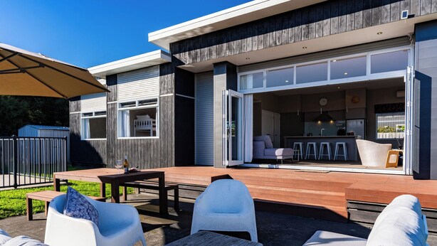A modern house with a patio area featuring outdoor furniture, a wooden deck, and a kitchen visible through the open sliding doors.