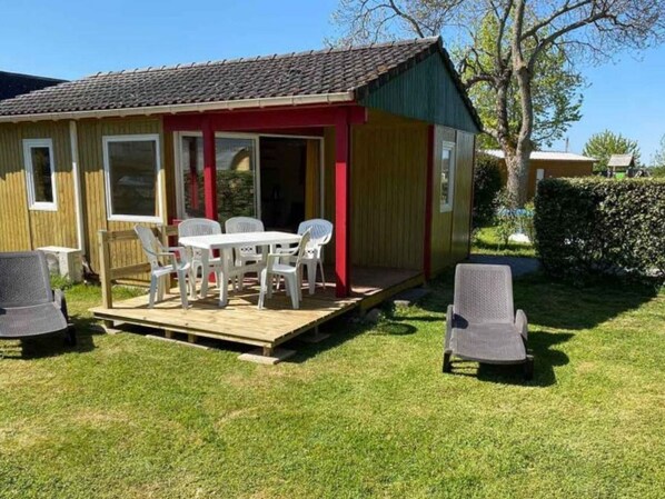 A small, single-story wooden house with a red door and windows, a wooden deck, and a table with four chairs.