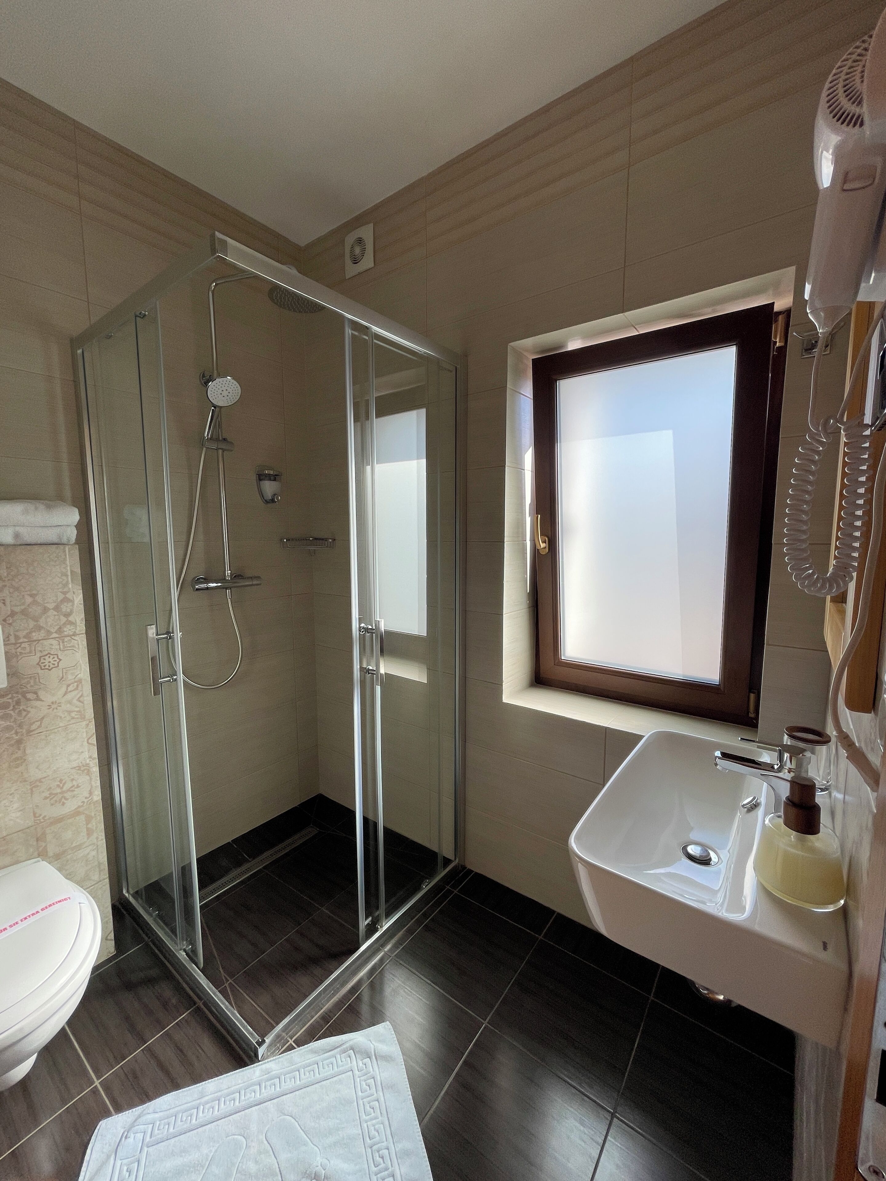 Un baño moderno con ducha envuelta en vidrio, un lavabo blanco y una ventana sobre el lavabo.