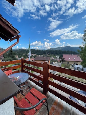Un balcon avec une table et des chaises, donnant sur un village avec ses maisons et une chaîne de montagnes.