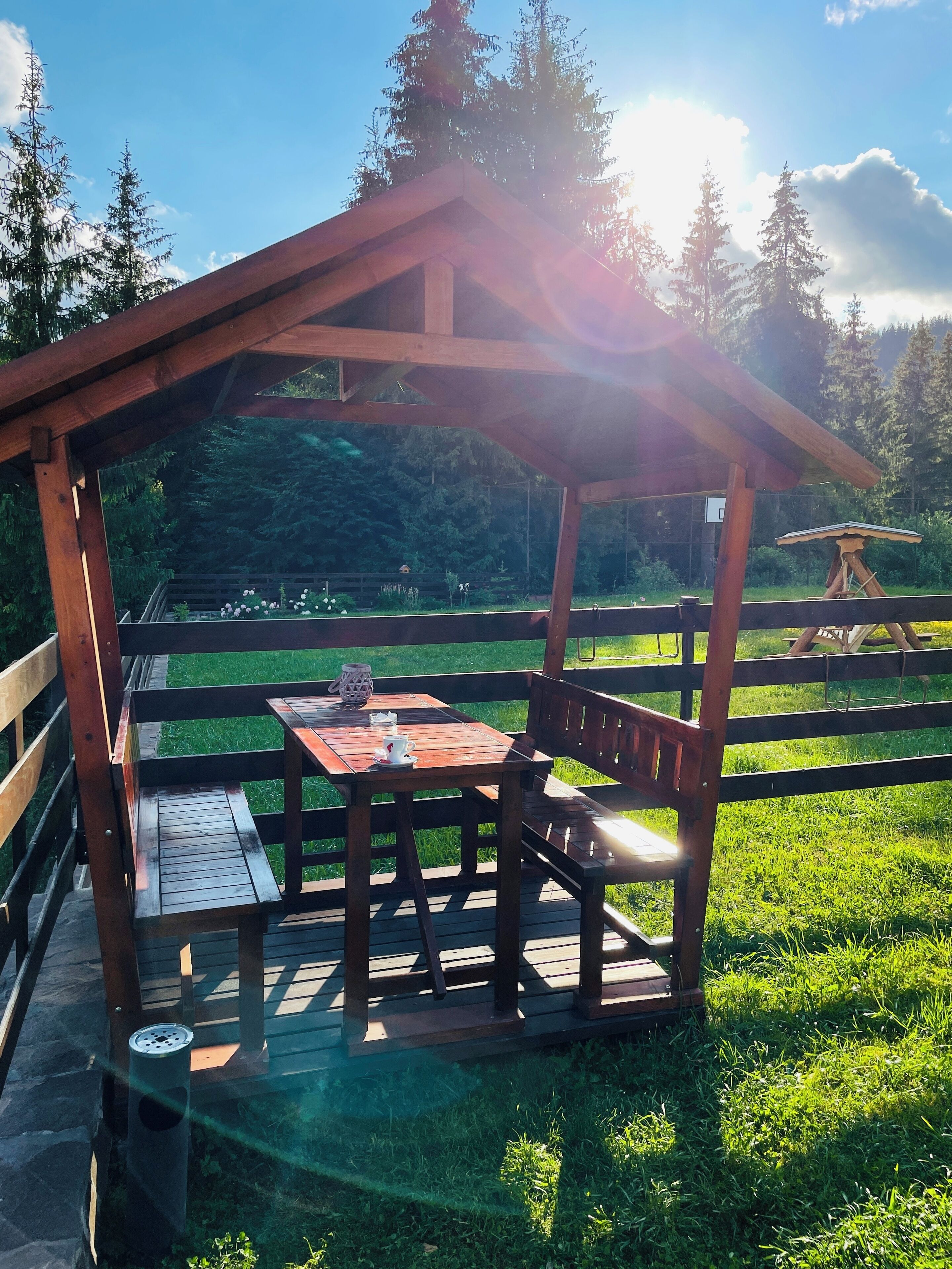 Ein hölzernes Gartenhaus mit Tisch und Bänken, umgeben von einer Rasenfläche und Bäumen.