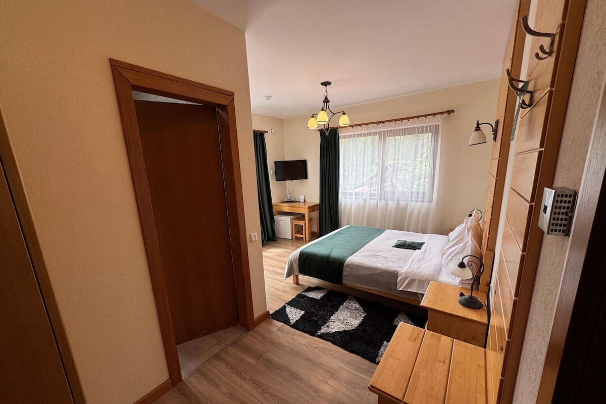 A bedroom with a bed, a desk, a chair, a TV, and a window with curtains.