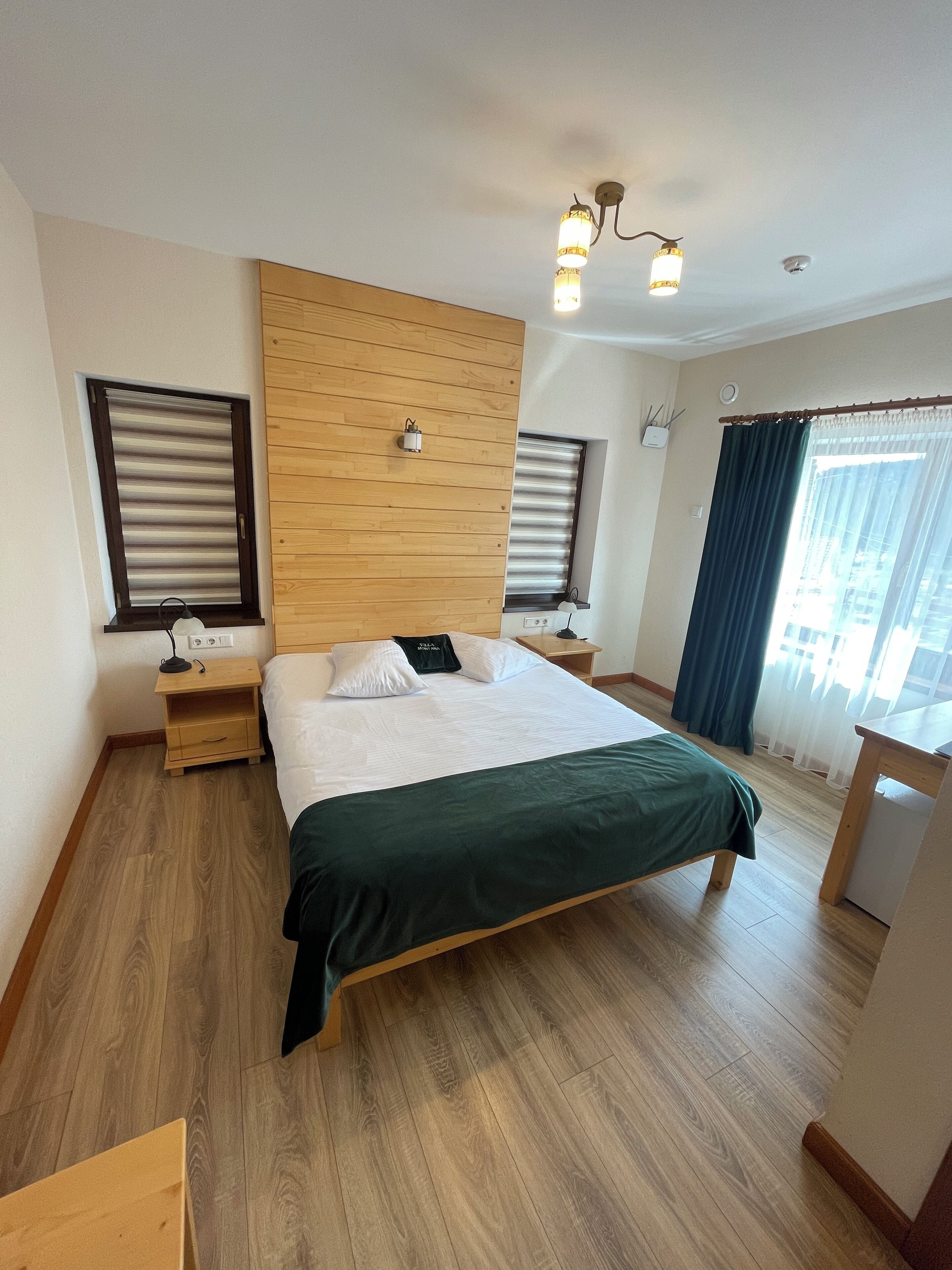 Camera d'albergo moderna con una testiera in legno, un letto grande con copriletto verde, due comodini con lampade, un tavolino notte in legno e vista sulla città attraverso tende leggere.