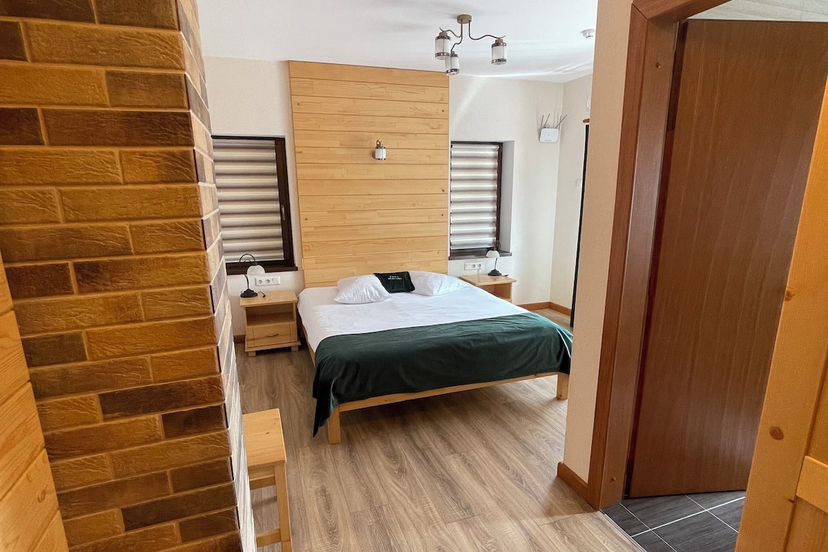 A modern bedroom with a wooden headboard, a bed with a green bedspread, a nightstand, a lamp, and a window with blinds.