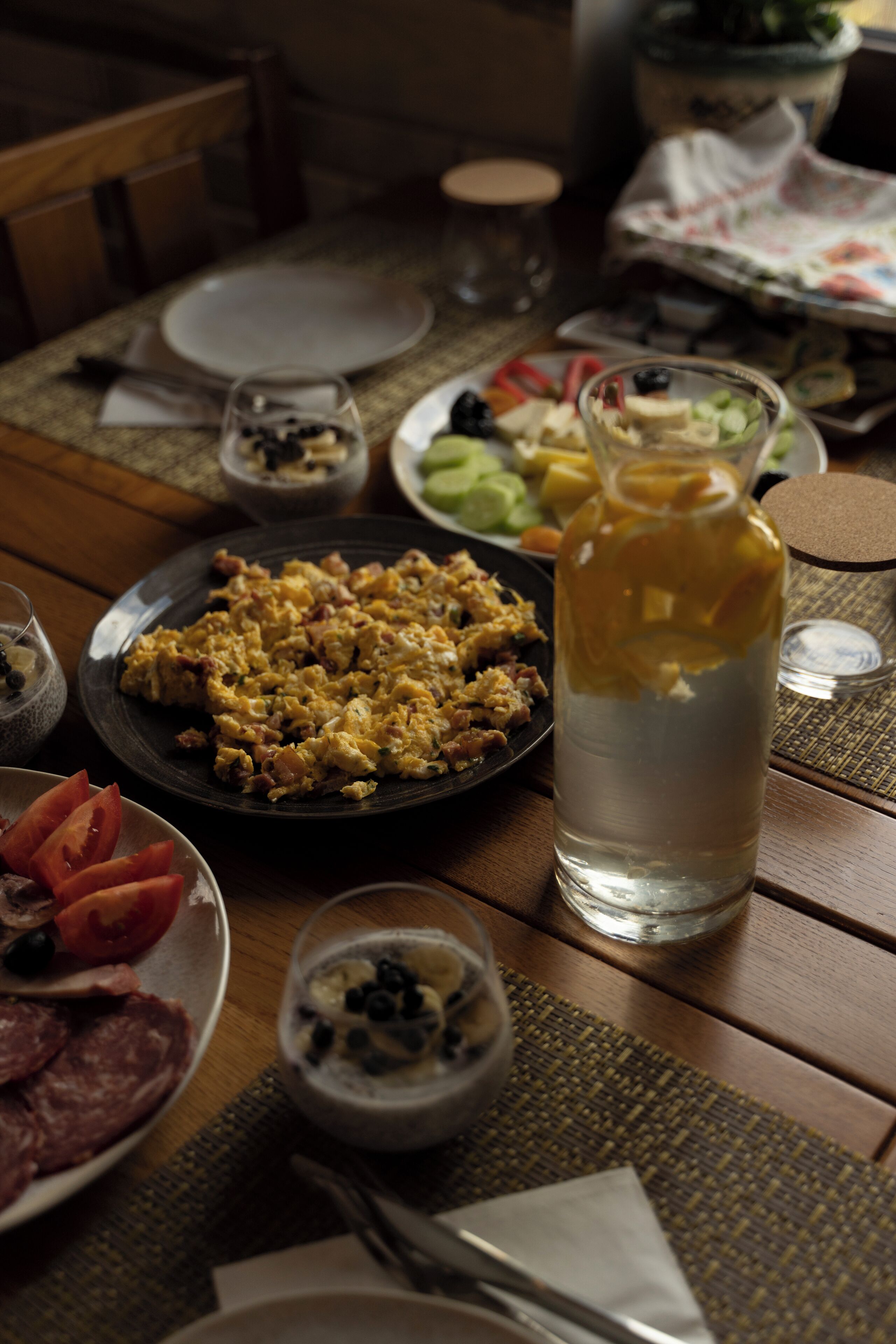 Una mesa de comedor con diversos platos, entre ellos huevos revueltos, una jarrón de agua y un bol de fruta.